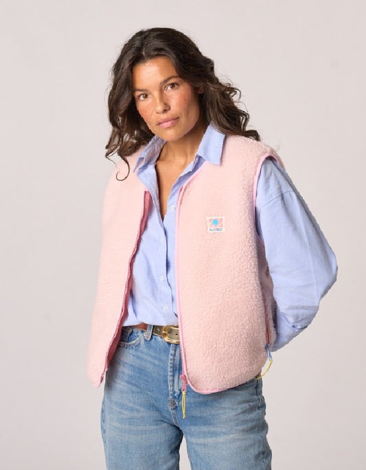 A woman wearing a light pink sherpa fleece vest over a light blue button-down shirt and blue jeans, photographed against a plain light gray studio background.