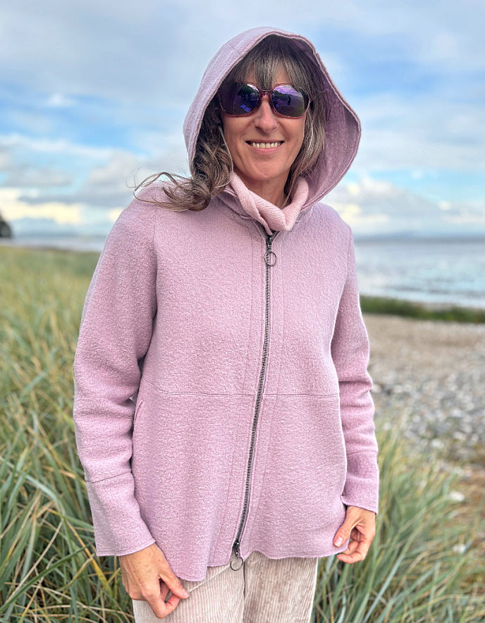 Person wearing a pink boiled wool hoodie jacket on a beach with grass and water in the background