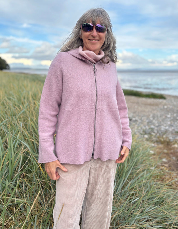 Person wearing a pink boiled wool hoodie jacket on a beach with grass and water in the background