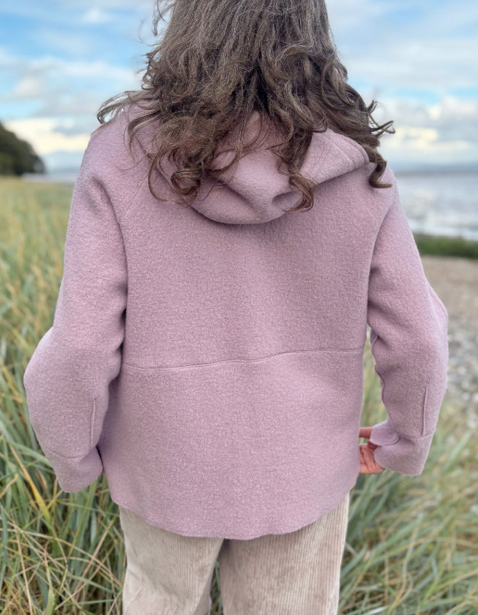 Person wearing a pink boiled wool hoodie jacket on a beach with grass and water in the background