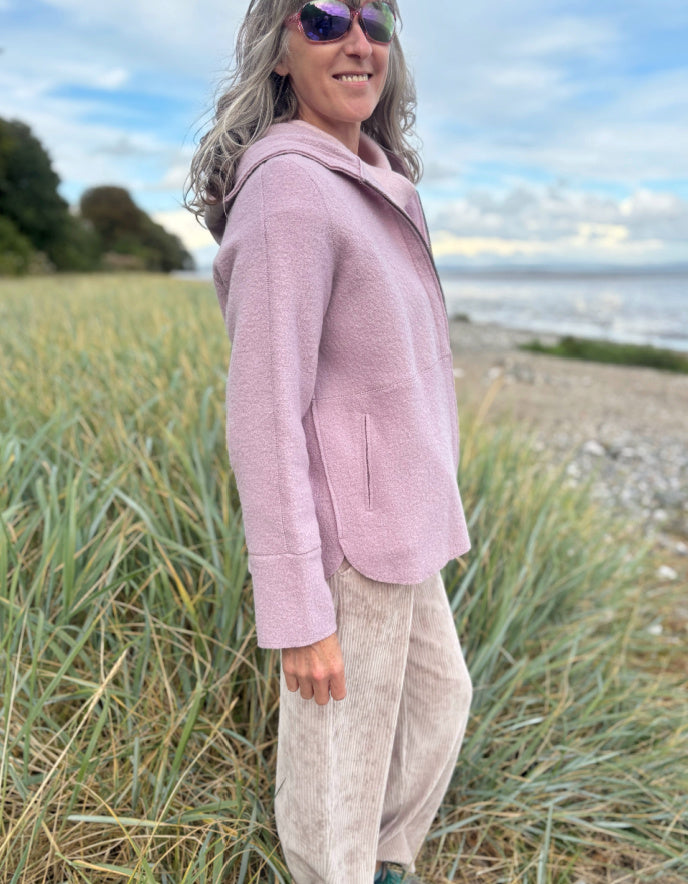 Person wearing a pink boiled wool hoodie jacket on a beach with grass and water in the background