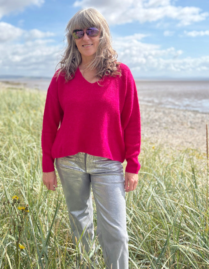 Woman in a pink v neck sweater and sunglasses standing in grass near a beach.