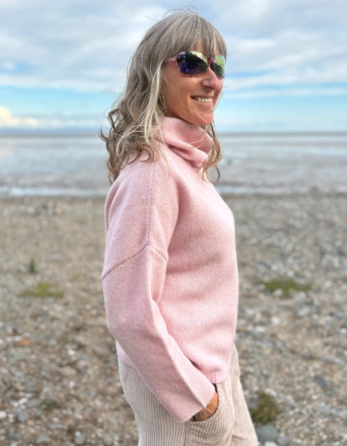 Woman wearing a pink sweater on a beach with a cloudy sky.