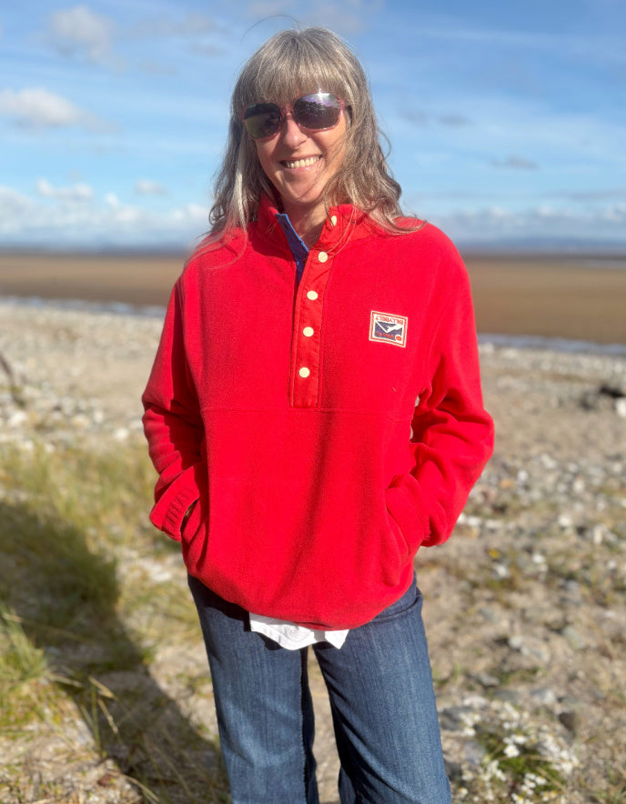 Person wearing a red jacket with a logo, sunglasses, and jeans standing on a rocky beach.
