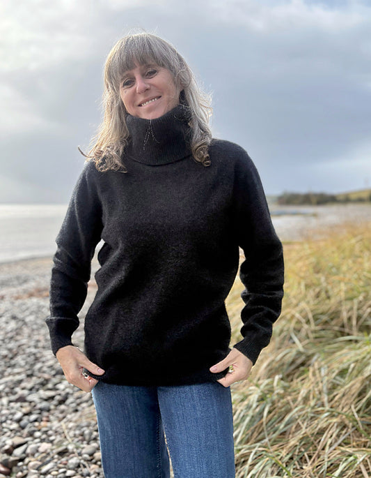 Person wearing a black turtleneck sweater standing on a beach with grass and water in the background.