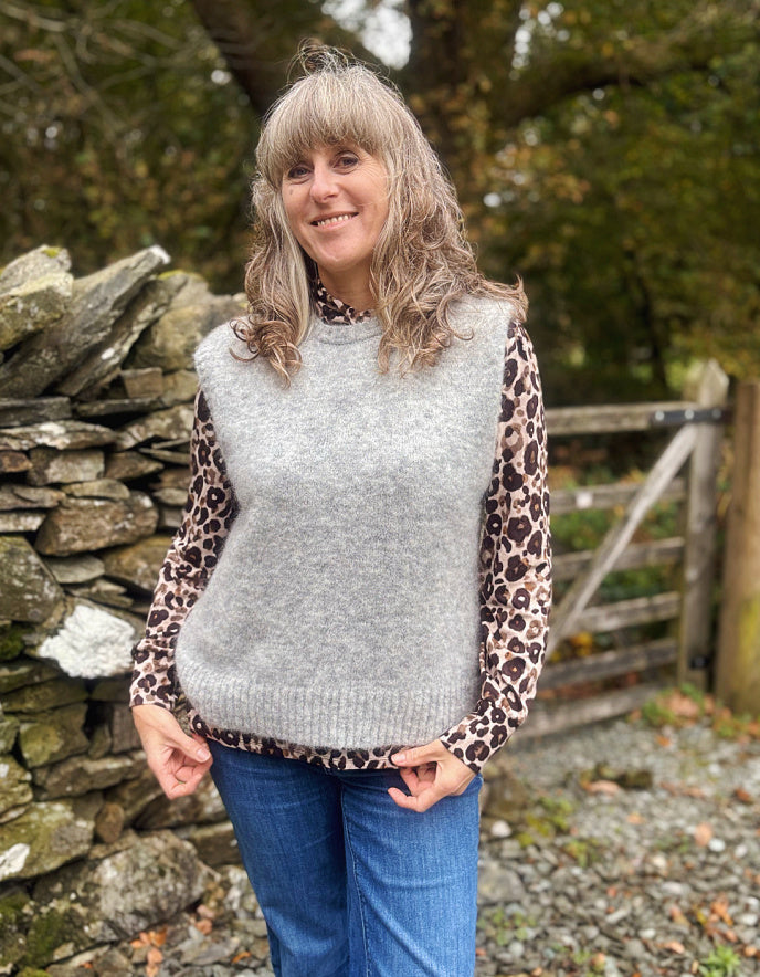 Woman wearing a light gray sleeveless sweater over a leopard print shirt with blue jeans, standing in front of a stone wall and wooden gate.