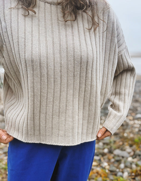 A person wearing a ribbed knit sweater in a light oatmeal beige color, paired with bright blue pants, standing outdoors on a pebble-strewn shore.