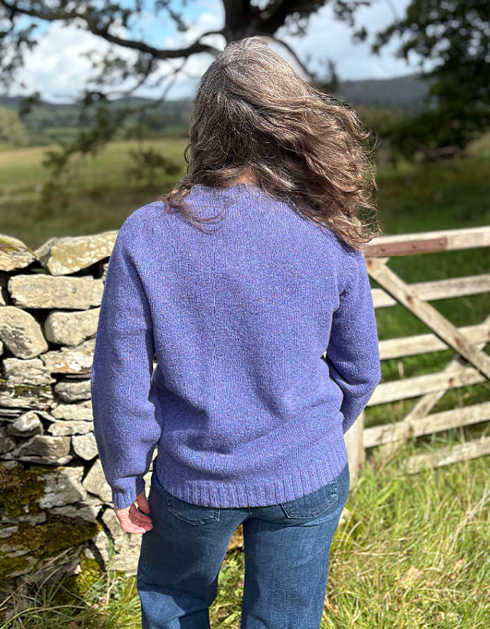 Person wearing a purple donegal tweed cardigan standing in a field with a stone wall and wooden gate in the background.