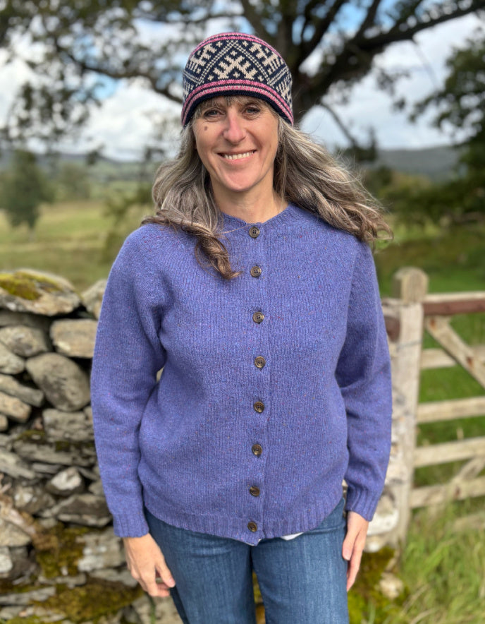 Woman wearing a purple donegal tweed knitted cardigan and patterned hat standing in a natural setting with trees and a stone wall.