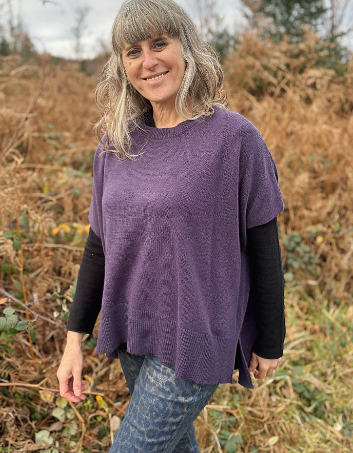Woman wearing a purple sweater standing in a natural setting with dry grass and trees.