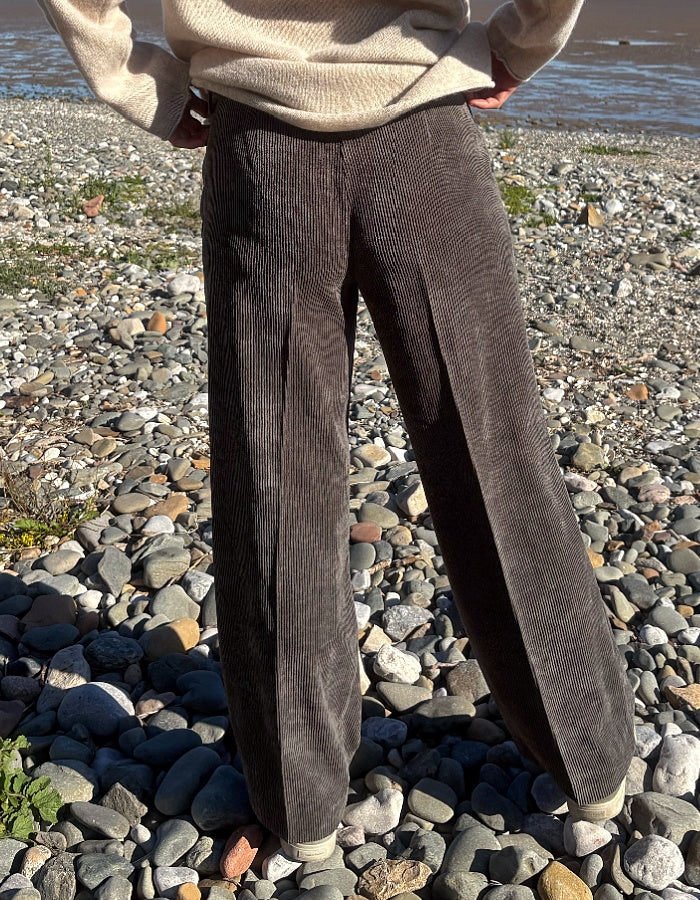 A person standing on a pebble beach wearing dark brown corduroy wide-leg trousers and a beige knit sweater, with water visible in the background.
