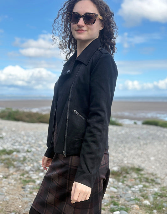A woman with curly hair wearing a black suede moto jacket and a brown plaid skirt, standing on a rocky beach with a blue sky in the background.