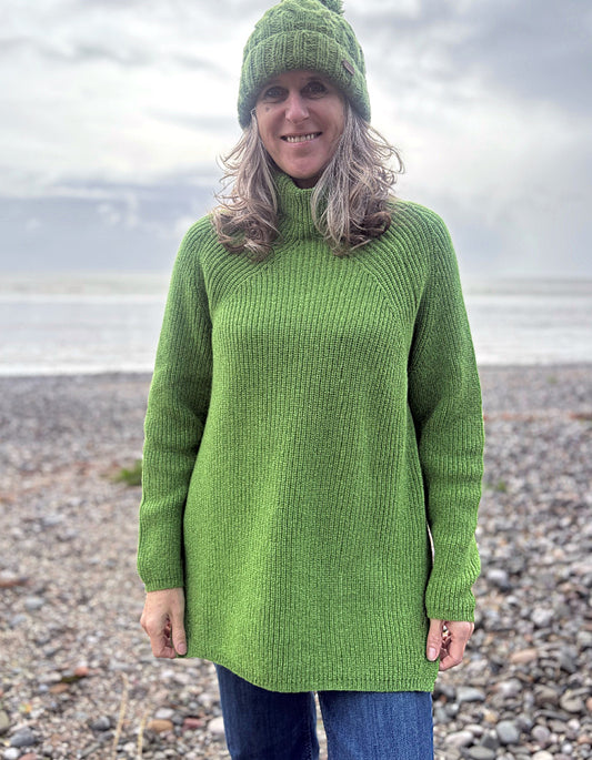 Person wearing a green knitted sweater and hat standing on a pebbly beach.