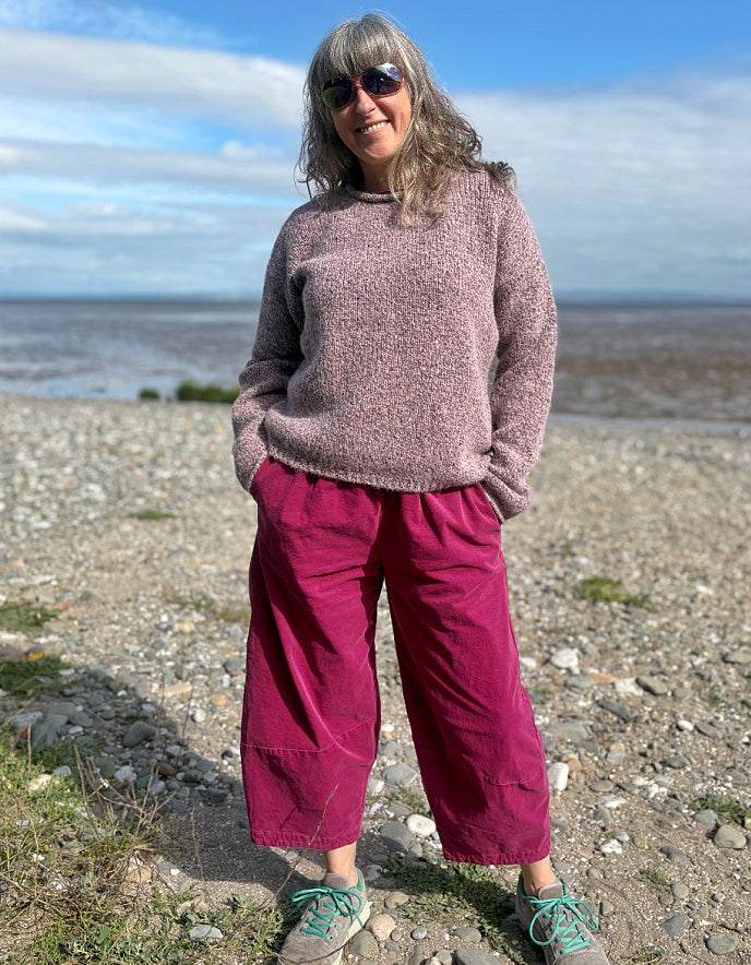 Person wearing a pink sweater and purple pants standing on a pebbly beach with a blue sky.