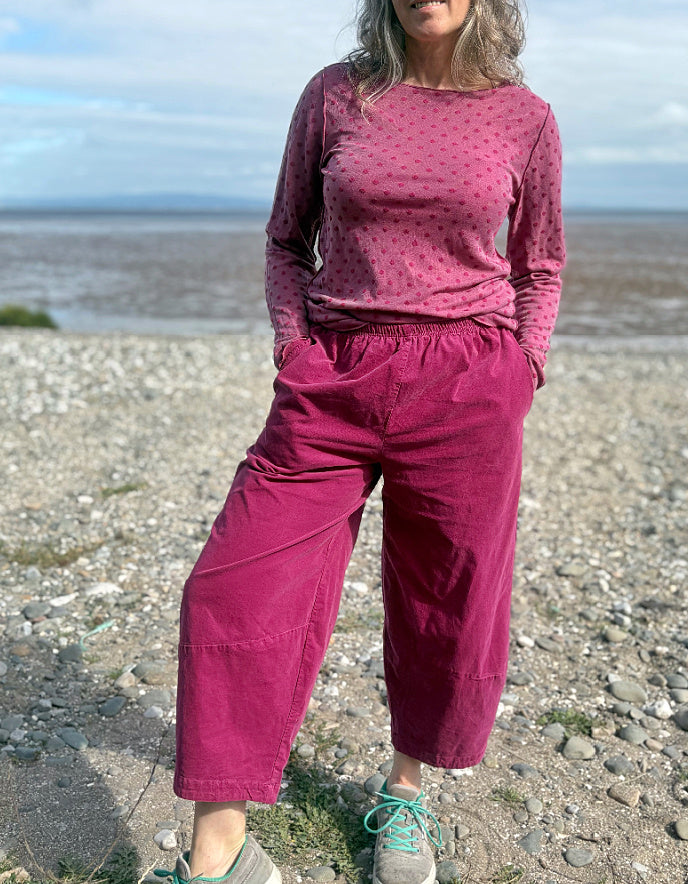Woman in pink needlecord lantern trousers and a pink pink spotty top standing on a pebbly beach with ocean and sky in the background