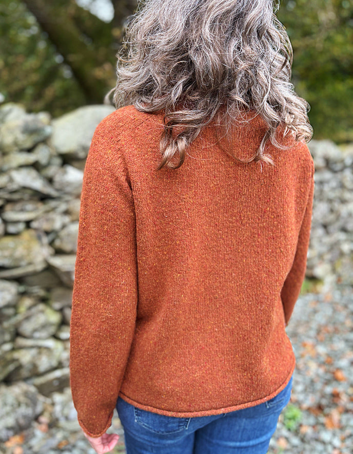 Person wearing a rust-colored sweater with a stone wall and greenery in the background