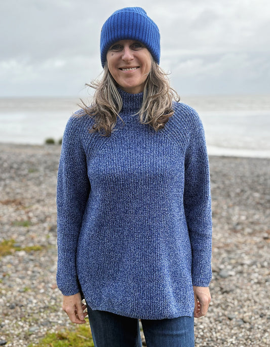 Person wearing a blue knit sweater and beanie standing on a pebbly beach.