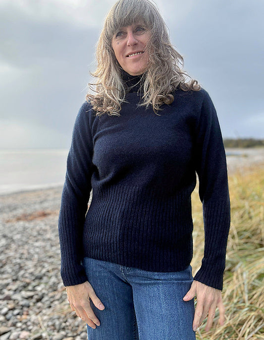 Person wearing a navy sweater and blue jeans standing on a beach.