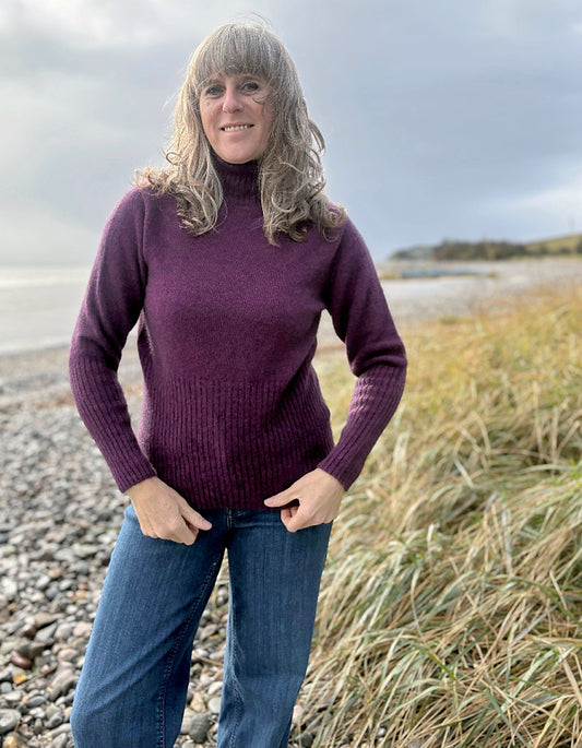 Woman wearing a purple sweater and blue jeans standing on a beach.