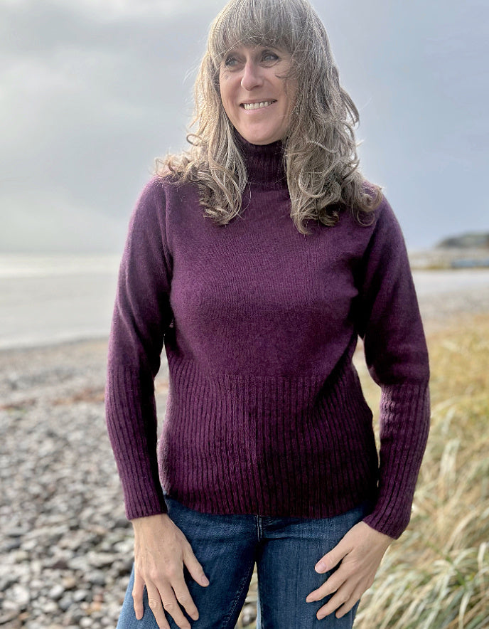 Woman wearing a purple sweater standing on a beach with a cloudy sky.