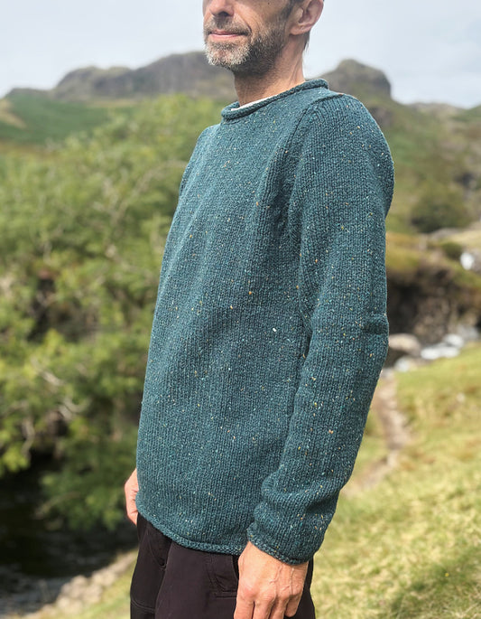 A man wearing a textured, speckled teal-green knit sweater, standing outdoors in a grassy, rocky landscape.
