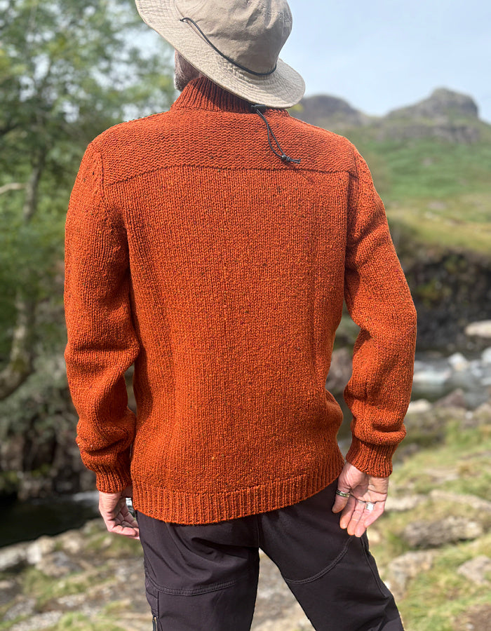 A man wearing a textured rust-orange knit sweater, a beige wide-brim hat, and dark trousers, photographed from behind in an outdoor mountain landscape.