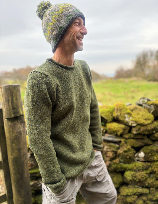 Man wearing a green sweater and knit hat standing in a natural setting with a stone wall and grass.