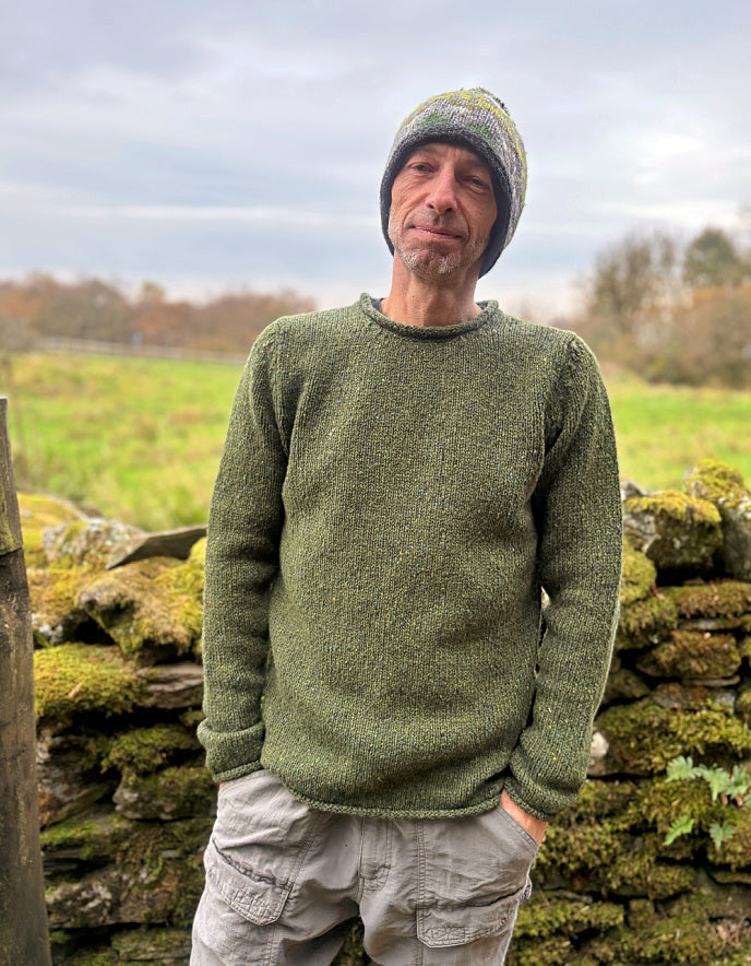Man wearing a green knitted sweater and beanie standing in front of a stone wall with grass and trees in the background.