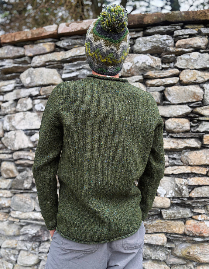 Person wearing a green knitted sweater and colorful beanie standing against a stone wall.