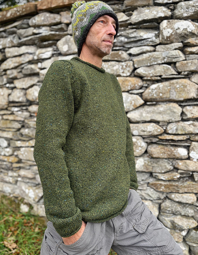 Man wearing a green knitted sweater and beanie standing against a stone wall.