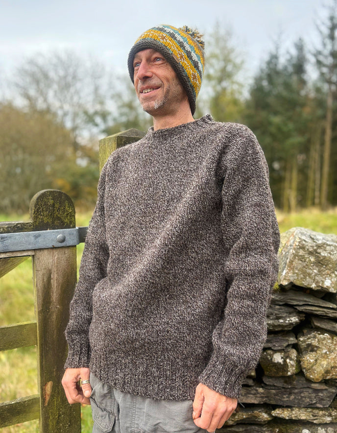 Man wearing a knitted sweater and colorful beanie standing by a wooden gate in a rural setting.