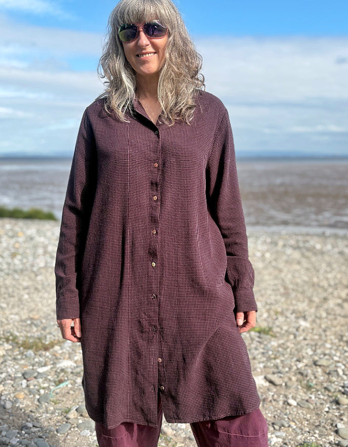 Person wearing a midi length Burgundy shirt dress garment on a pebbly beach with a blue sky.