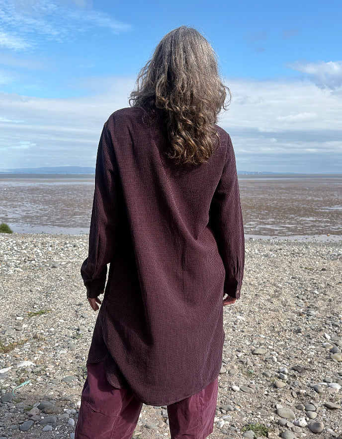 Person wearing a midi length Burgundy shirt dress garment on a pebbly beach with a blue sky.