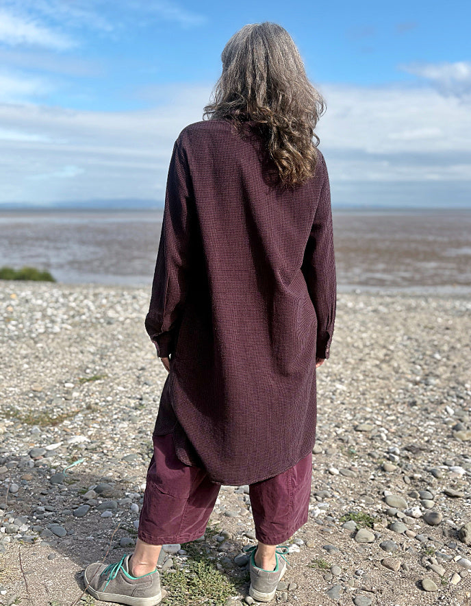 Person wearing a midi length Burgundy shirt dress garment on a pebbly beach with a blue sky.