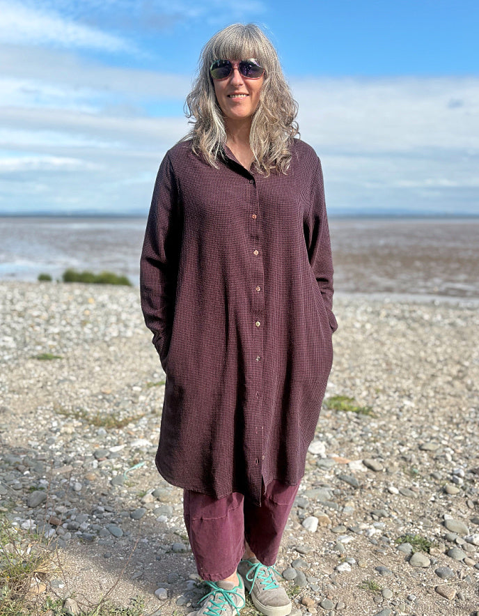 Person wearing a midi length Burgundy shirt dress garment on a pebbly beach with a blue sky.
