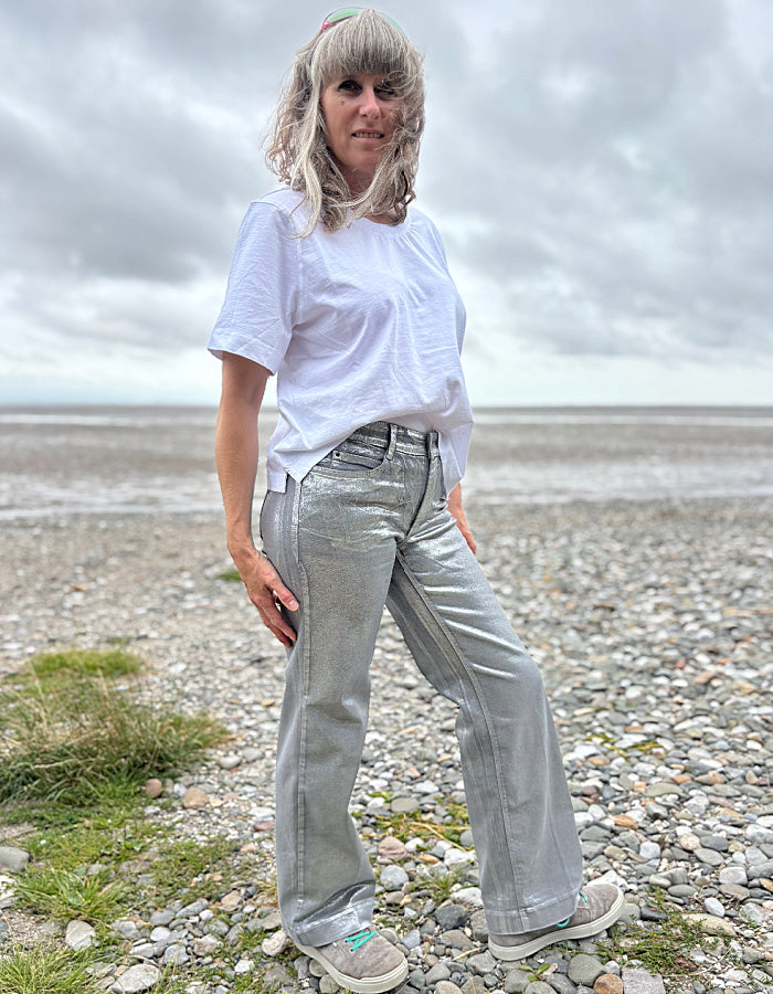 Person wearing a white t-shirt and light silver wide leg jeans standing on a pebbly beach with a cloudy sky.