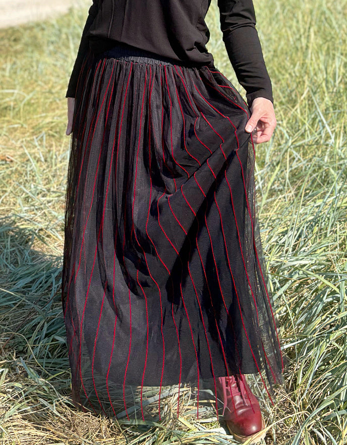 Woman standing in a field wearing a black long-sleeve top and a long, maxi length dark skirt with red stripes.