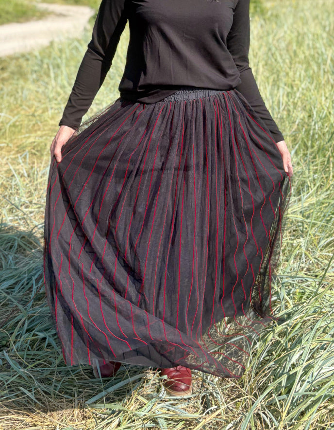 Woman standing in a field wearing a black long-sleeve top and a long, maxi length dark skirt with red stripes.