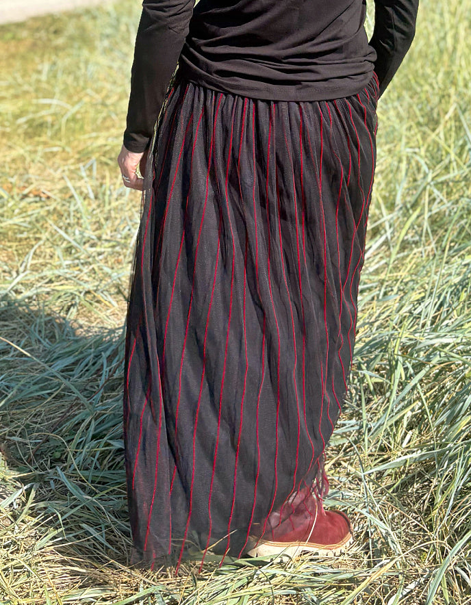 Woman standing in a field wearing a black long-sleeve top and a long, maxi length dark skirt with red stripes.