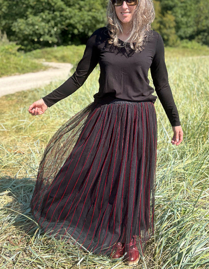 Woman standing in a field wearing a black long-sleeve top and a long, maxi length dark skirt with red stripes.