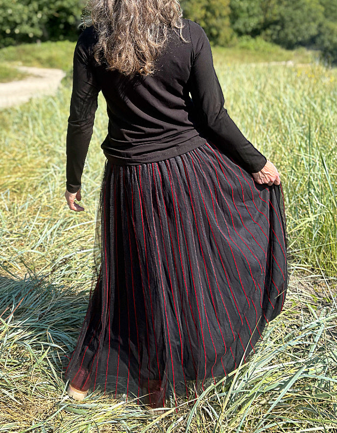 Woman standing in a field wearing a black long-sleeve top and a long, maxi length dark skirt with red stripes.