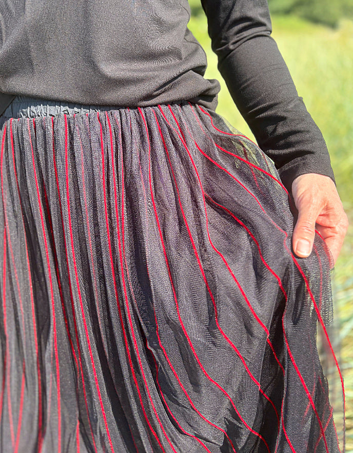 Woman standing in a field wearing a black long-sleeve top and a long, maxi length dark skirt with red stripes.