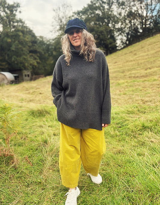 A woman standing outdoors on a grassy hill wearing an oversized dark gray turtleneck sweater, mustard yellow pants, white sneakers, and a dark blue cap.