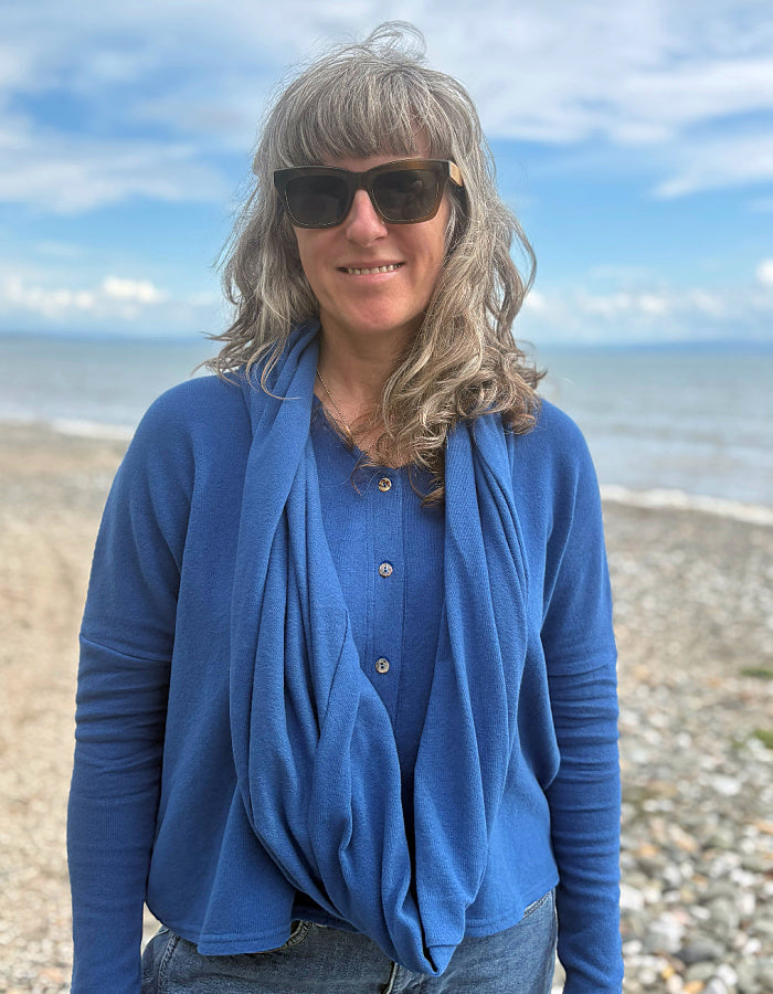 A woman with gray hair and sunglasses stands on a pebble beach wearing a blue long-sleeve top and a draped blue scarf, with the ocean and blue sky in the background.