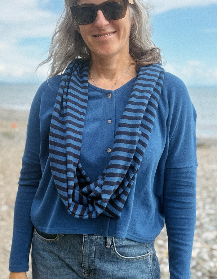 A woman wearing a blue long-sleeve henley shirt and a navy and blue striped infinity scarf, standing on a beach with the ocean and sky in the background.