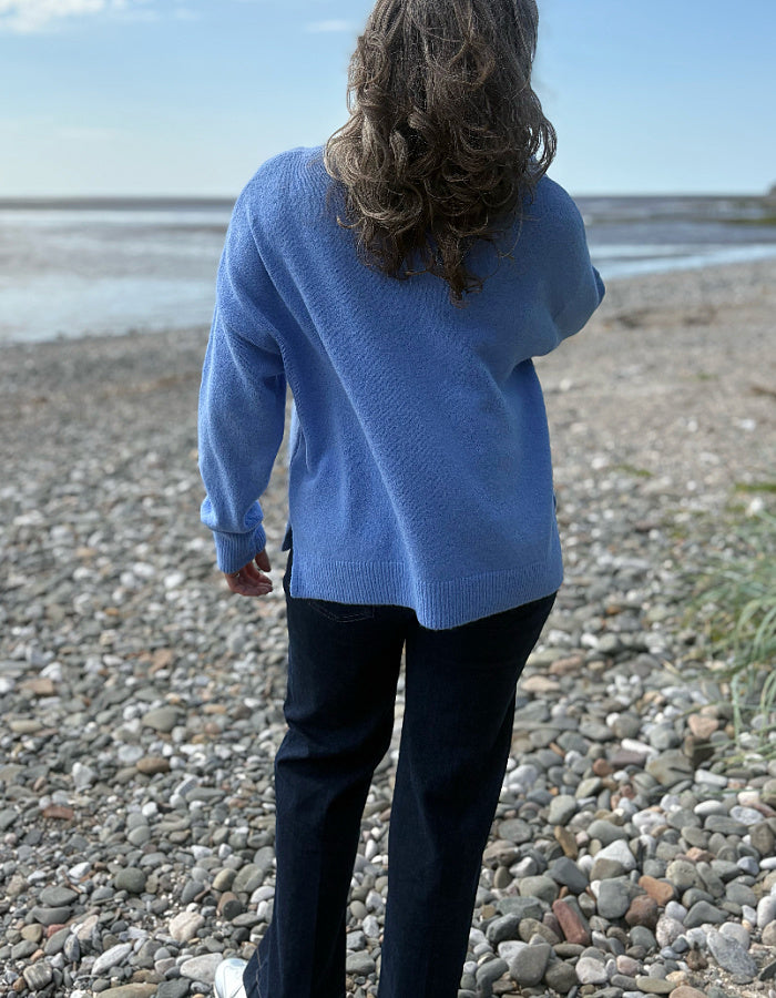 Person wearing a blue sweater standing on a pebbly beach with water and sky in the background
