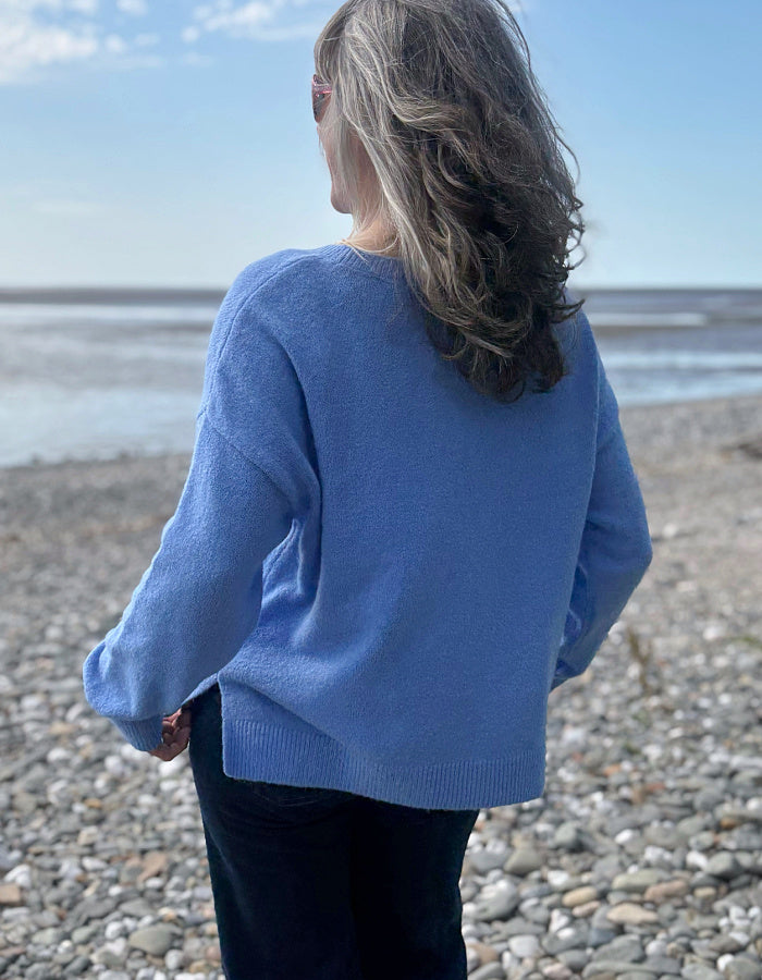 Person wearing a blue sweater standing on a pebbly beach with ocean and sky in the background
