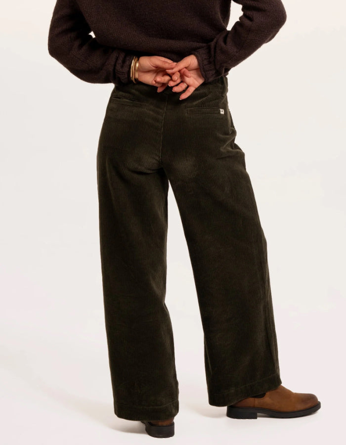 A person wearing dark olive green wide-leg corduroy pants, styled with a dark brown sweater and brown leather shoes, shown from the waist down on a white studio background.