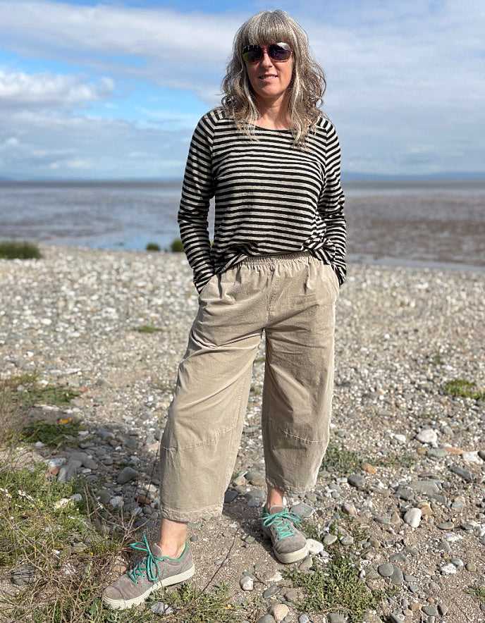 Person standing on a pebbly beach wearing a striped shirt and beige pants.