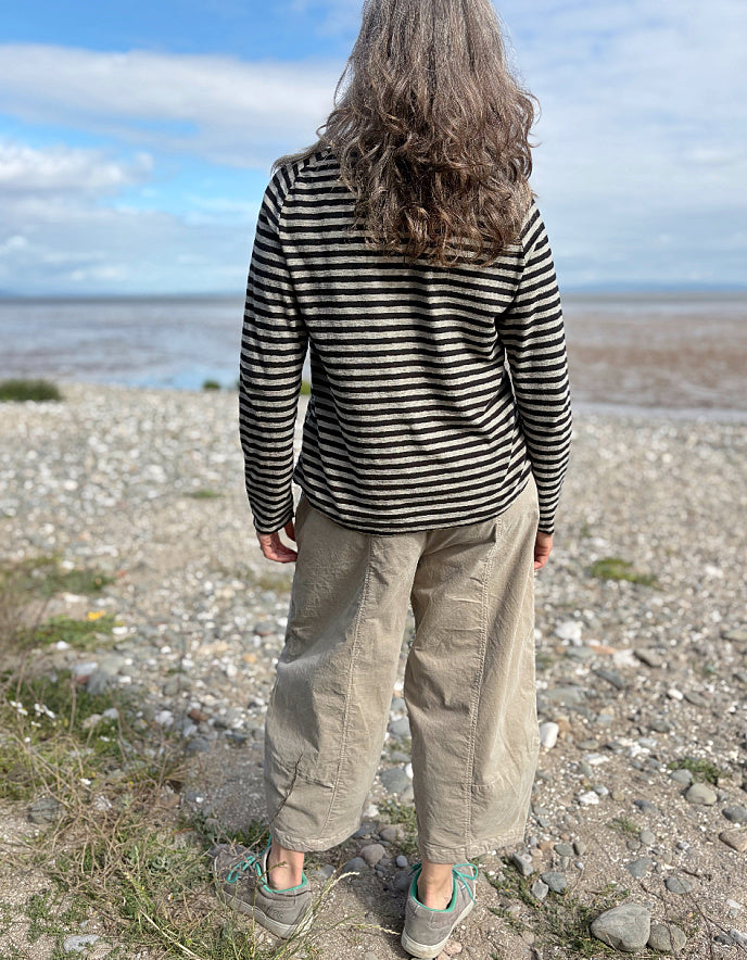 Person wearing a striped shirt and beige pants standing on a pebbly beach with water and sky in the background.